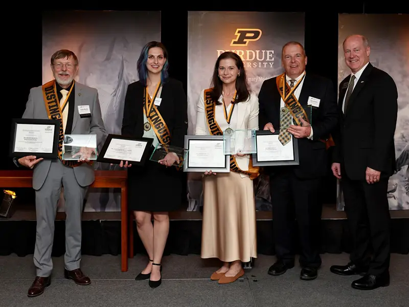 Five people pose at a Purdue University awards ceremony, holding certificates and wearing recognition sashes.